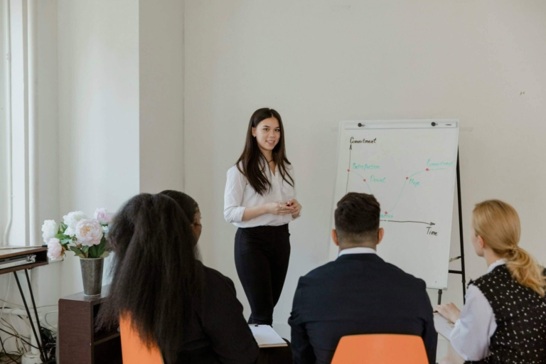 Femme leader animant une session de coaching collectif en leadership féminin devant tableau blanc, avec équipe mixte attentive, illustrant coaching d'équipe et intelligence collective.