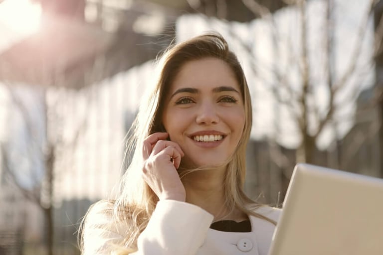 Jeune femme dirigeante souriante au téléphone avec tablette numérique, en extérieur devant un bâtiment moderne, symbolisant le leadership féminin confiant et la connectivité professionnelle.