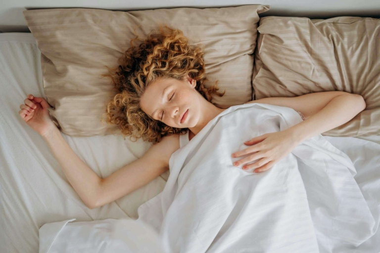 Femme aux cheveux bouclés dormant d'un sommeil profond et réparateur, visage serein sur oreiller beige, symbole de qualité du sommeil grâce à la sophrologie : endormissement facile, cycles respectés, gestion réveils nocturnes.